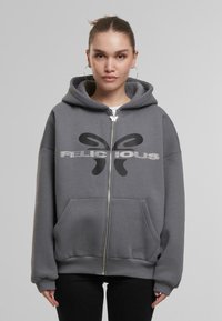 Grauer Reißverschluss-Hoodie mit einer Fronttasche, versehen mit einem großen schwarzen "FELICIOUS"-Logo und einem dezenten glänzenden Designelement. Weicher Stofftextur.