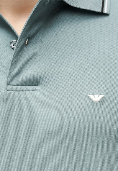 Φωτεινό γαλάζιο-πράσινο πόλο μπλουζάκι με γιακά, ένα κουμπί κουμπωμένο και ένα μικρό λευκό κεντημένο λογότυπο στο στήθος.