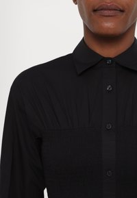 Musthave mustaga shirt klassikalise kraega, millel on smockitud tekstuur kehapiirkonnas ja sile kangas kaelaosas.