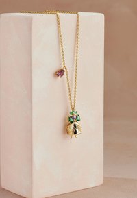 Guldhalsband med ett hänge i form av en klänning prydd med gröna och lila ädelstenar, visad på en blekrosa kub.