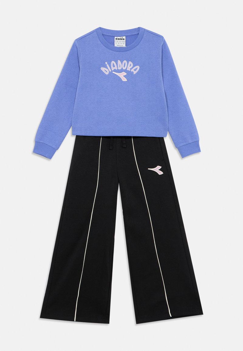 Felpa blu con logo "DIADORA" in rosa, abbinata a pantaloni larghi neri con piping laterale bianco e accentuazione del logo rosa. In misto cotone.