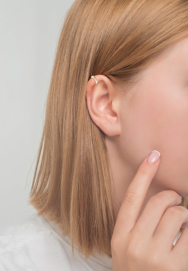 Boucle d'oreille en or au design incurvé avec trois petites pierres, portée sur une coiffure lisse et châtain clair sur fond neutre.