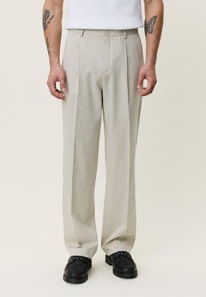 Mann trägt beige plissierte Hose, schwarze Leder-Loafers und weißes Hemd, zeigt Tätowierungen auf den Unterarmen vor einfarbigem Hintergrund.