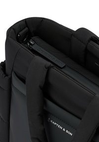 Svart vadderad väska med slät utsida, två sorters tygtexturer, dragkedja upptill och handtag. Har präglat logotyp på sidan.