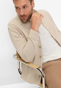Beige button-up shirt with a smooth texture, featuring a collar and a single chest pocket, worn over a white T-shirt and brown pants.