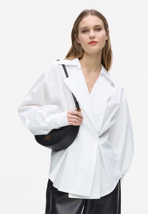 Jonge vrouw draagt een oversized witte blazer en zwarte leren broek, met een zwarte halfmaanvormige handtas met gouden beslag over haar schouder.