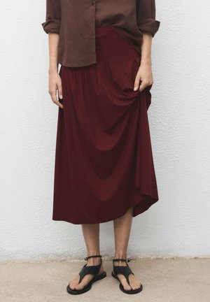 Burgundy midi skirt in a flowing fabric with draped details, paired with black flat sandals and a brown loose button-up shirt.
