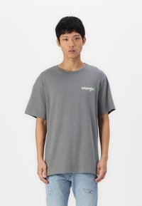 Szara bawełniana koszulka z okrągłym dekoltem i krótkimi rękawami. Na lewej stronie znajduje się małe, zielone logo "Wrangler". Noszona z jasnoniebieskimi, zniszczonymi dżinsami.