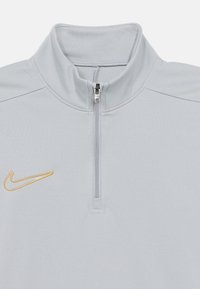 Ανοιχτό γκρι πουλόβερ Nike με γιακά μισού φερμουάρ, που διαθέτει χρυσό κεντημένο λογότυπο στο αριστερό στήθος και υφασμάτινη υφή.