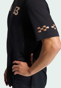 Musta t-paita, jossa on suuri "63" vaaleammassa sävyssä ja metallinhohtoisia pronssinvärisiä ruutukuvioita hihassa, valmistettu pehmeästä puuvillamateriaalista.