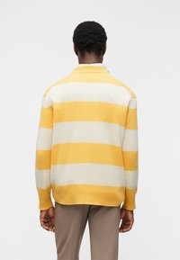 Άτομ ο που φοράει κίτρινο και λευκό πουλόβερ με φαρδιές ρίγες και γκρι παντελόνι, στέκεται γυρισμένο προς τα πίσω μπροστά σε απλό φόντο.