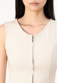 Beige ärmlös klänning med rund halsringning och metallisk dragkedja framtill, tillverkad av texturerat tyg med subtila sömdetaljer.