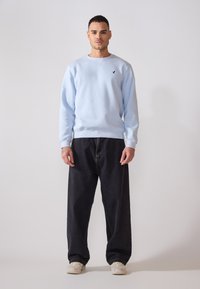 Ljusblå sweatshirt med rund halsringning och svart logotyp, i kombination med löst sittande mörka jeans och ljusa sneakers.