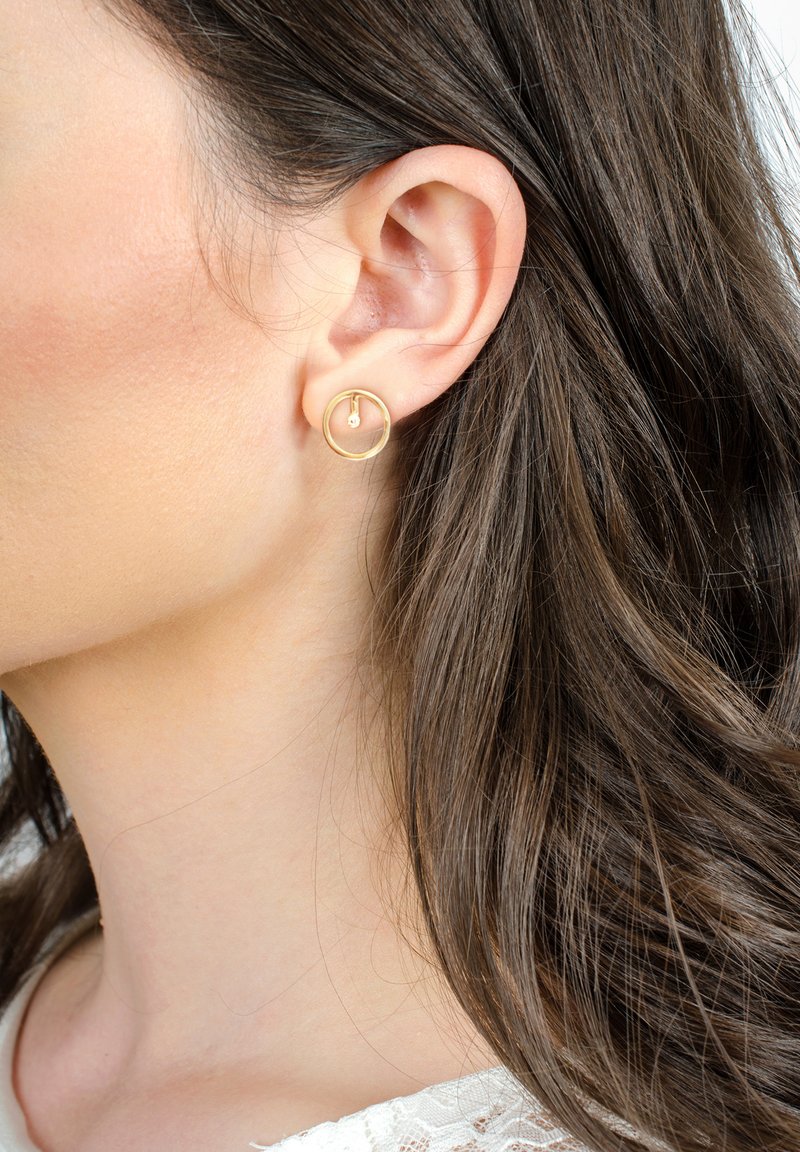Orecchino a cerchio dorato con un design circolare e un piccolo accento a goccia, indossato sull'orecchio sinistro, accompagnato da capelli lisci e castano scuro.