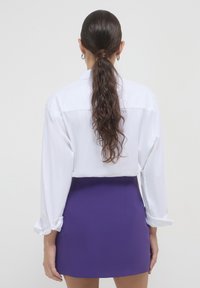 Camicia bianca a maniche lunghe con polsini risvoltabili abbinata a una gonna corta e aderente di colore viola. Tessuto liscio senza motivi o decorazioni visibili.