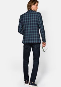 Marynarka w kratę w ciemnym niebieskim i zielonym kolorze, dopasowany krój, zestawiona z ciemnymi dżinsami. Tkanina jest teksturowana; widok z tyłu podkreśla fason i dopasowanie.