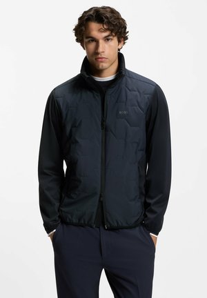 Giovane uomo con capelli ricci che indossa una giacca imbottita nera con zip e pantaloni scuri, in piedi con le mani in tasca, espressione neutra.