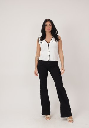 Vrouw staand, gekleed in een mouwloos wit vest met knopen en zwarte uitlopende broek, met lichtbeige open-toe hakken, tegen een effen achtergrond.