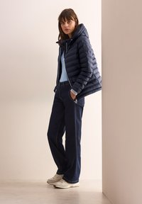 Giacca puffer blu navy con cappuccio, top azzurro chiaro, pantaloni in denim scuro e sneakers chiare. Tessuto liscio e tasche con zip sui pantaloni.