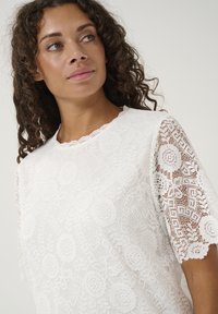 White lace top featuring intricate floral patterns, scalloped neckline, and short sleeves with sheer accents. Soft texture and lightweight design.