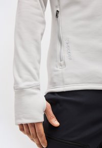 Κοντινό πλάνο ενός ατόμου που φοράει ένα ανοιχτό γκρι μπουφάν με πλαϊνή τσέπη με φερμουάρ και μανσέτα με τρύπα για τον αντίχειρα, σε συνδυασμό με μαύρο παντελόνι με τσέπη με φερμουάρ.