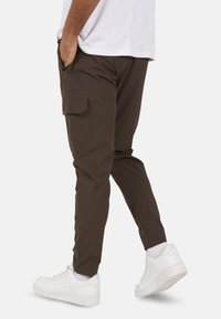 Brązowe spodnie typu cargo z bocznymi kieszeniami i zwężanym krojem, w połączeniu z białymi trampkami. Tkanina wydaje się lekka i gładka.