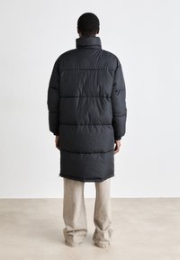 Long black puffer coat with horizontal stitching, high collar, and side slits, paired with light beige wide-leg trousers.