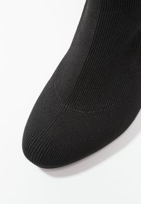 Gros plan sur un bout de chaussure en tissu tricoté côtelé noir avec une forme arrondie sur fond blanc.
