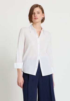 Donna con capelli corti che indossa una camicia bianca con bottoni e maniche arrotolate, e pantaloni larghi blu scuro, su uno sfondo semplice.