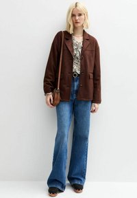 Giacca marrone, testurizzata, con colletto a scollo a V, abbinata a una blusa floreale e jeans blu a gamba larga. Gli accessori includono una borsa a spalla marrone.
