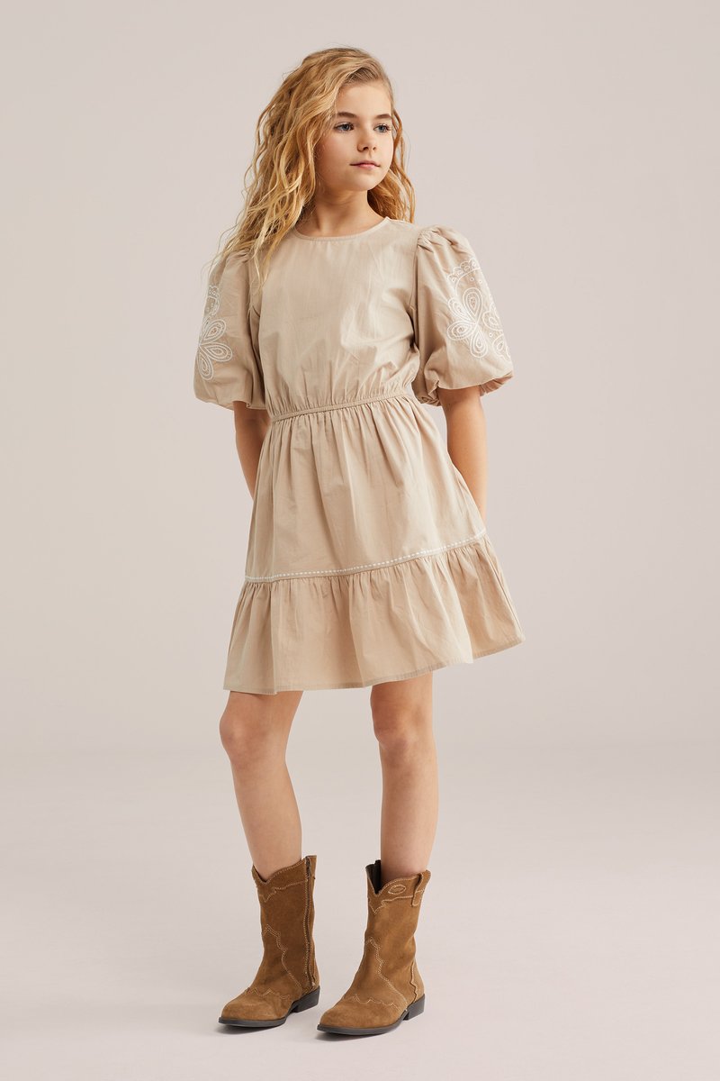 Jauna mergina su ilgais šviesiais plaukais, vilkinti kreminės spalvos su pūstomis rankovėmis su baltomis siuvinėjimo detalėmis su rudais zomšiniais bato vidurio blauzdos aukščio bato, stovinti pasitikinti savimi.