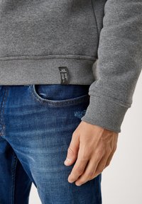Felpa grigia con polsini e orlo a coste, dotata di un'etichetta nera in basso. Abbinata a jeans di denim blu. Dettaglio ravvicinato della mano sui jeans.