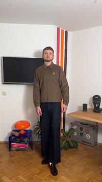 Καφέ μπουφάν με φερμουάρ και γιακά, σκούρο τζιν παντελόνι φαρδύ και σκούρα παπούτσια. Το δωμάτιο έχει τηλεόραση, πορτοκαλί λάμπα και φυτά.