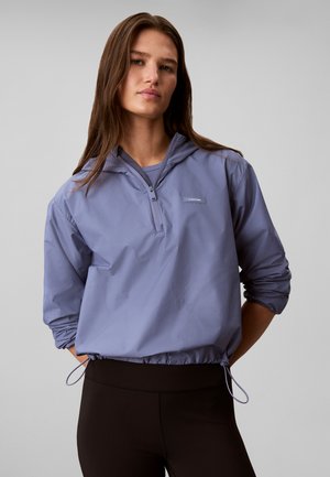 Femme aux longs cheveux bruns portant un sweat à capuche violet clair à fermeture éclair mi-hauteur et des leggings noirs sur un fond gris uni.