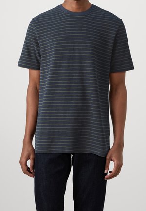 Homme portant un t-shirt bleu marine à manches courtes avec de fines rayures horizontales grises et un pantalon sombre, debout devant un fond uni.