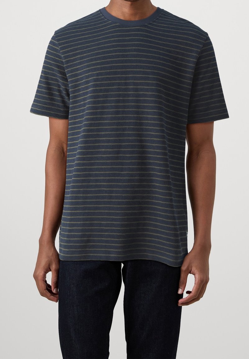 Homme portant un t-shirt bleu marine à manches courtes avec de fines rayures horizontales grises et un pantalon sombre, debout devant un fond uni.