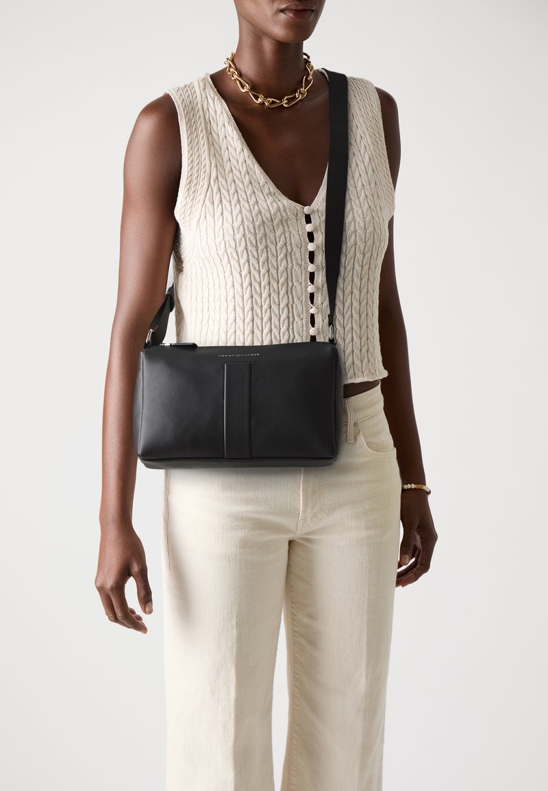 Sac à bandoulière en cuir noir de forme rectangulaire, avec une texture lisse et un détail sur le panneau avant. Bandoulière réglable pour plus de polyvalence.