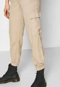 Beige Cargo-Hosen mit elastischen Bündchen und Seitentaschen, kombiniert mit schwarzen Schnürkampfstiefeln mit glänzender Sohle.