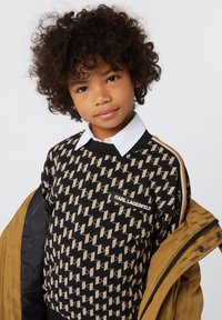 Sweatshirt noir avec un motif géométrique beige, col ras du cou, et patch de marque. Porté avec une veste beige, présentant une texture matelassée et des détails de design.