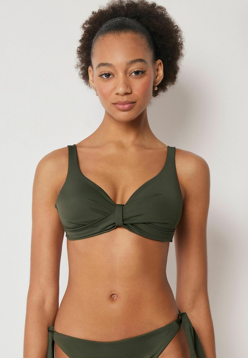 Jeune femme aux cheveux bouclés naturels porte un haut de bikini vert olive et un bas assorti à nouer sur les côtés, sur un fond uni.