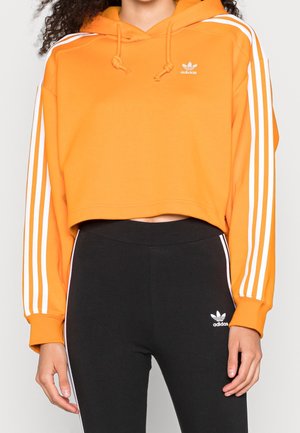 Vrouw draagt een korte oranje Adidas hoodie met witte strepen en zwarte Adidas leggings met witte strepen aan de zijkanten.