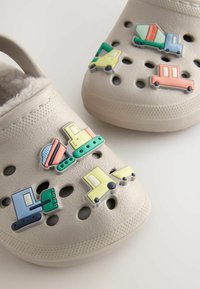 Zoccoli beige chiaro con numerosi fori di ventilazione, dotati di colorati charm in gomma a forma di veicolo nei colori blu, verde, giallo e rosa.