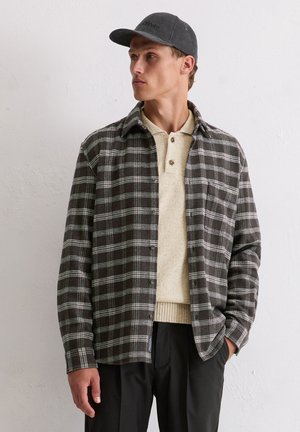 Uomo che indossa una camicia a quadri bianco e nero sopra un polo in maglia beige, pantaloni neri e un berretto grigio scuro, in piedi contro un muro bianco testurizzato.