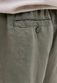 Gros plan sur la poche d'un pantalon vert olive avec un bouton et un tissu beige au-dessus de la ceinture.