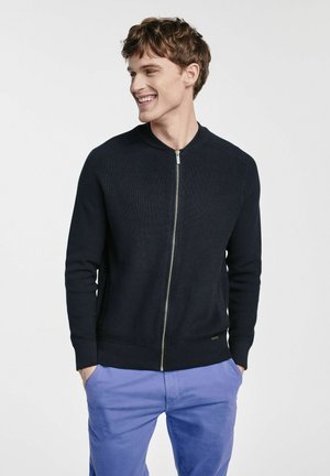 Granatowy sweter rozpinany na zamek o rypsowym wzorze, okrągłym dekolcie i metalowym detailu zamka błyskawicznego. Noszony z jasnofioletowymi spodniami.