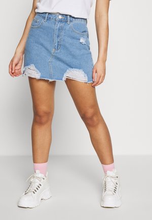 Kobieta ubrana w zniszczoną niebieską jeansową mini spódnicę, białe masywne sneakersy i różowe skarpetki, stojąca na tle gładkiej białej ściany.