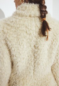 Cremefarvet faux fur jakke med en tekstureret, fluffy overflade og en bred krave. Har en synlig søm langs ryggen.