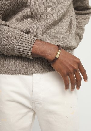 Persona de piel oscura que lleva un suéter gris de punto y pantalones blancos, con una pulsera de cuero trenzado marrón con un cierre dorado en la muñeca.
