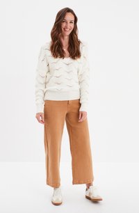 Maglione bianco lavorato a maglia con un motivo a onda, abbinato a pantaloni a gamba larga beige e sneaker bianche con dettagli marroni. Colori neutri, vestibilità rilassata.