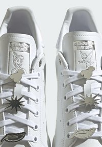 White leather Adidas Stan Smith sneakers featuring silver accents, character artwork on tongue, and metallic charms on laces in star and feather shapes.